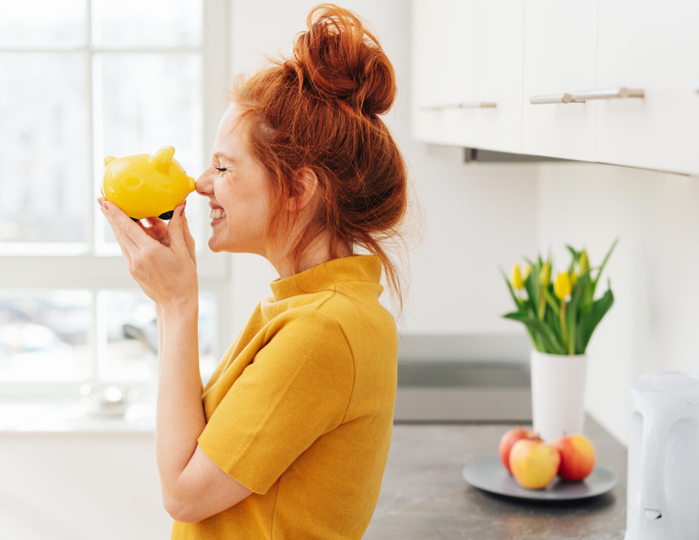 AdobeStock_257863957 Smiling red-haired woman playing with yellow piggy bank in her hands, viewed from the side in bright room interior. Money savings concept