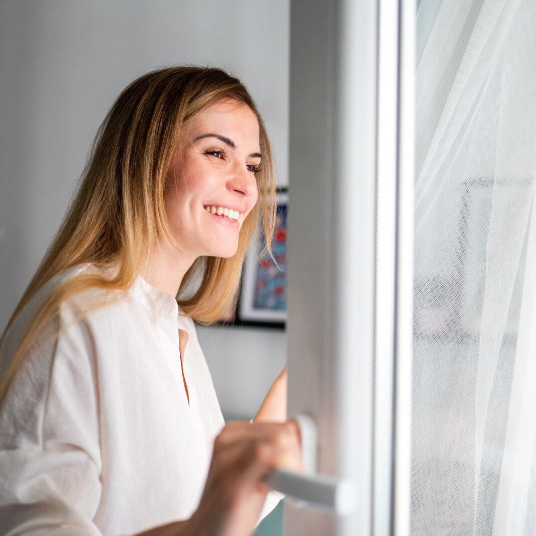 AdobeStock_377893241 Beautiful smiling young woman opening window at the morning, fresh air,
